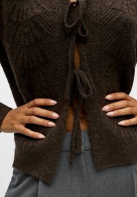 Pull en tricot marron avec un tissu texturé, doté d'une fermeture à nouer à l'avant et d'un design ouvert, porté sur un pantalon gris, les mains posées sur les hanches.