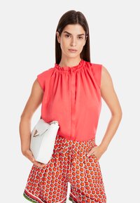 Coral sleeveless blouse with a ruffled neckline, paired with red shorts featuring a strawberry print. The model holds a white handbag.