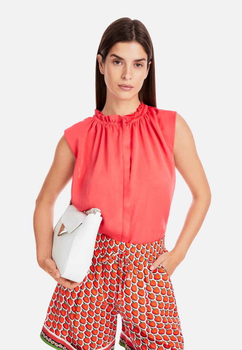 Coral sleeveless blouse with a ruffled neckline, paired with red shorts featuring a strawberry print. The model holds a white handbag.
