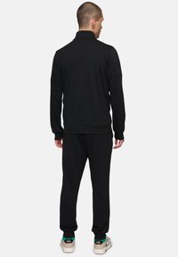 Homme de dos portant un survêtement noir et des chaussures FILA blanches avec des accents verts sur un fond clair.