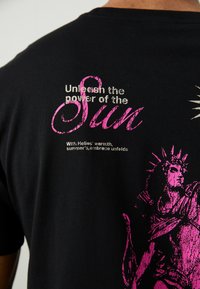 Schwarzes T-Shirt mit rosa und weißem Grafiktext sowie einer Statue-Illustration. Das Design betont sonnige Motive und fette Typografie.