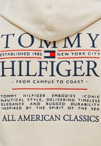Λευκό φούτερ με κουκούλα που διαθέτει μπλε και κόκκινη κεντητή γραφή, συμπεριλαμβανομένων των "TOMMY HILFIGER" και "ALL AMERICAN CLASSICS", με λεία υφή.