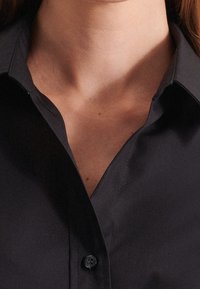 Black button-up shirt with a structured collar, smooth fabric, and a single button at the neck. Features a sharp, clean design.
