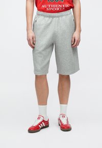 Άτομο που φοράει ανοιχτογκρι σορτς μέχρι το γόνατο, κόκκινη αθλητική μπλούζα, λευκές κάλτσες και κόκκινα αθλητικά παπούτσια Adidas με λευκές ρίγες.