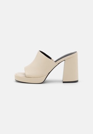 Beige High-Heel Offener Zehen-Mule mit breitem Riemen und Blockabsatz auf einem einfarbigen hellen Hintergrund.