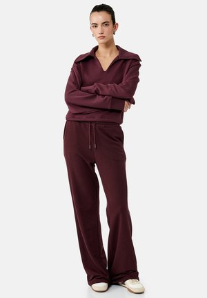 Kvinde med mørkt hår i en knold iført en burgunderfarvet V-hals sweatshirt og matchende vidde bukser, stående med armene krydset foran på en hvid baggrund.
