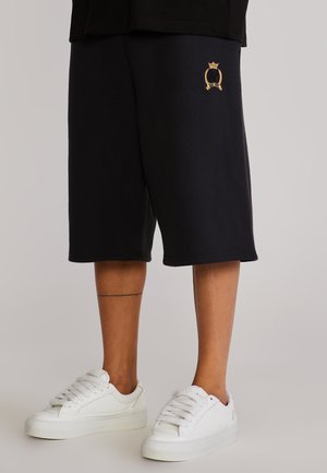 Person iført sorte shorts til knæene med et lille gyldent emblem på venstre lår og hvide lave sneakers.