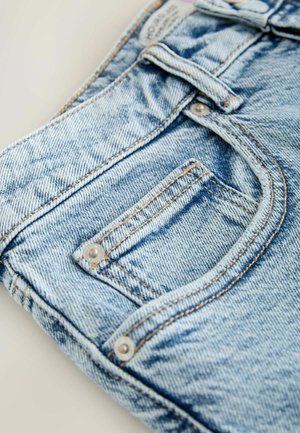 Detail svetlomodrých džínsy z denimu zobrazujúci prednú kapsu, pútko na opasok a oranžové prešívanie na rovnom, neutrálnom pozadí.