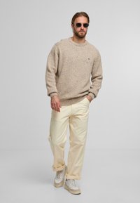 Beige gestrickter Pullover mit einem strukturierten Muster, cremefarbene weit geschnittene Hose und beige/graue Sneakers. Accessoire: runde Sonnenbrille.