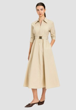 Donna con capelli scuri in un abito midi beige con cintura, cerniera frontale e maniche a tre quarti, che indossa scarpe marroni con tacco basso a punta, su sfondo grigio.