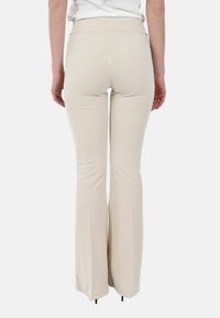 Donna che indossa pantaloni attillati beige vista da dietro, in piedi su sfondo bianco.