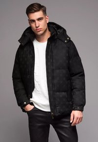 Svart puffjacka med en strukturerad mönstring, stor huva och guldfärgad detaljering. Bärs över en vit sweatshirt och svarta byxor.