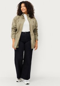 Khaki utilityjakke med ståkrave og lynlåslommer, parret med en hvid turtleneck og marineblå viddebukser, komplementeret af hvide sneakers.