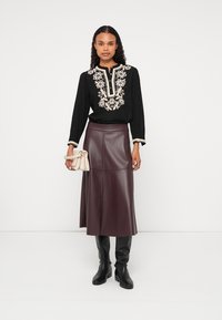 Čierna blúzka s kvetinovou výšivkou, skombinovaná s bordovou sukňou z umlej kože. Modelka má na sebe vysoké čierne čižmy a drží krémovú kabelku.