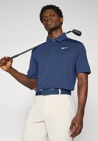 Granatowa koszulka polo golfowa z fakturowanym wzorem, z krótkim rękawem i logo Nike; w zestawie z jasnobeżowymi spodniami i dopasowanym niebieskim paskiem.