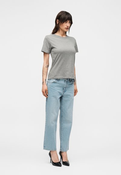 Γυναίκα με σκούρα μαλλιά φοράει απλό γκρι κοντομάνικο μπλουζάκι, ανοιχτό μπλε ίσια τζιν, μαύρα ψηλά τακούνια και δείχνει τατουάζ και στα δύο πήχεις.