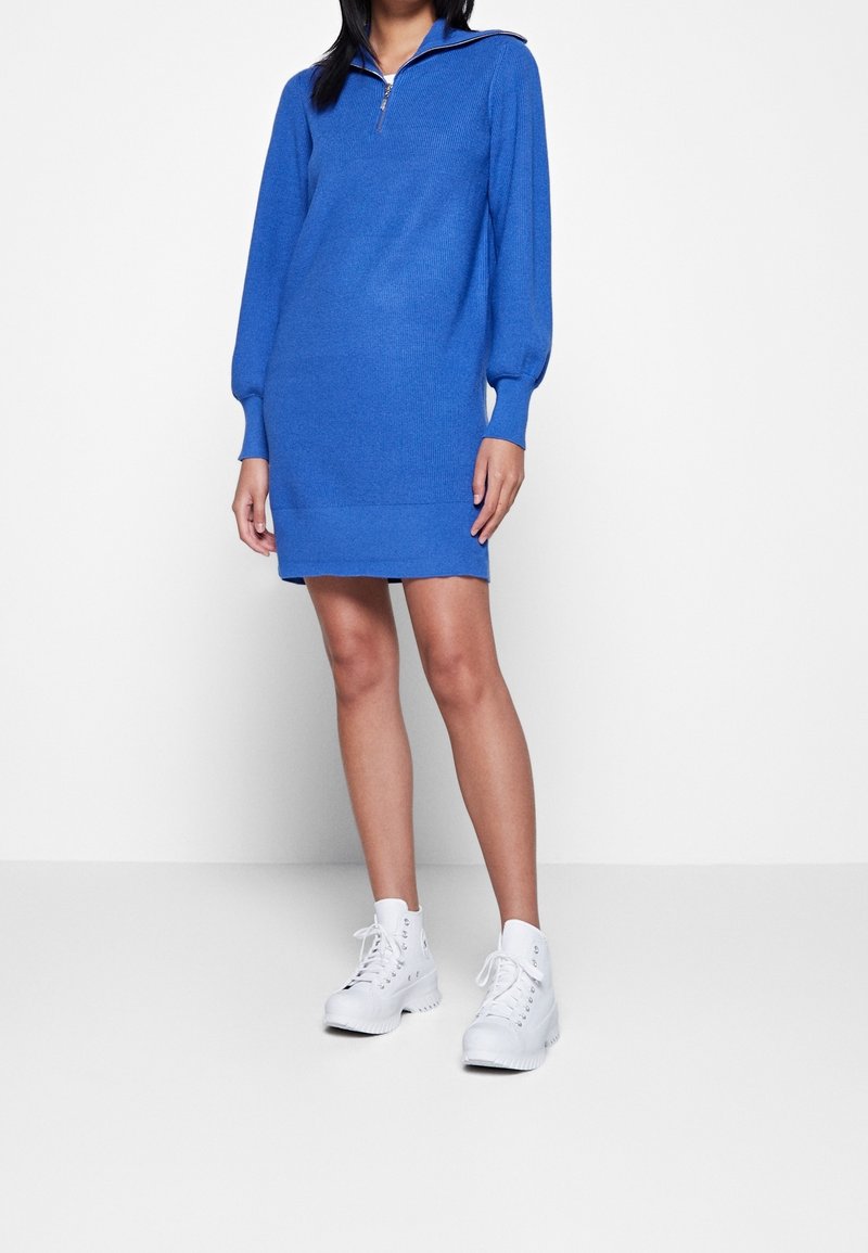 Robe en tricot bleu avec de longues manches bouffantes et un col zippé mi-hauteur, présentant une texture côtelée et une coupe droite, associée à des chaussures blanches.