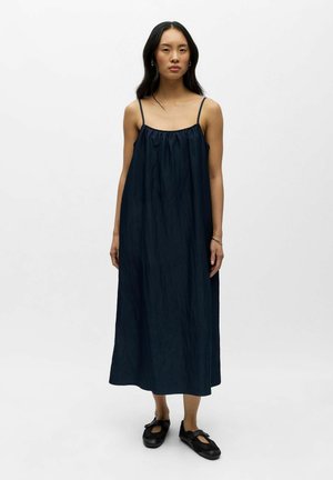 Donna in piedi che indossa un abito midi senza maniche di colore blu scuro con spalline sottili e scarpe nere stile Mary Jane su sfondo bianco.