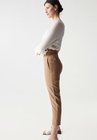 Brune skreddersyde bukser med folder, høj talje og slank pasform. Sættet er kombineret med en struktureret cremesweater og slidesandaler. Neutral baggrund.