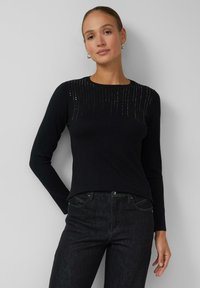Schwarzer Pullover mit langen Ärmeln, strukturiert mit einem Rippenmuster und dekorativen Verzierungen am Ausschnitt. Kombiniert mit dunklen Jeans.