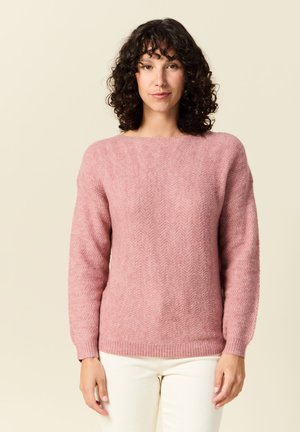 Pull rose à motif chevron avec un col rond et des manches longues, fabriqué en matériau texturé doux, associé à un pantalon blanc.