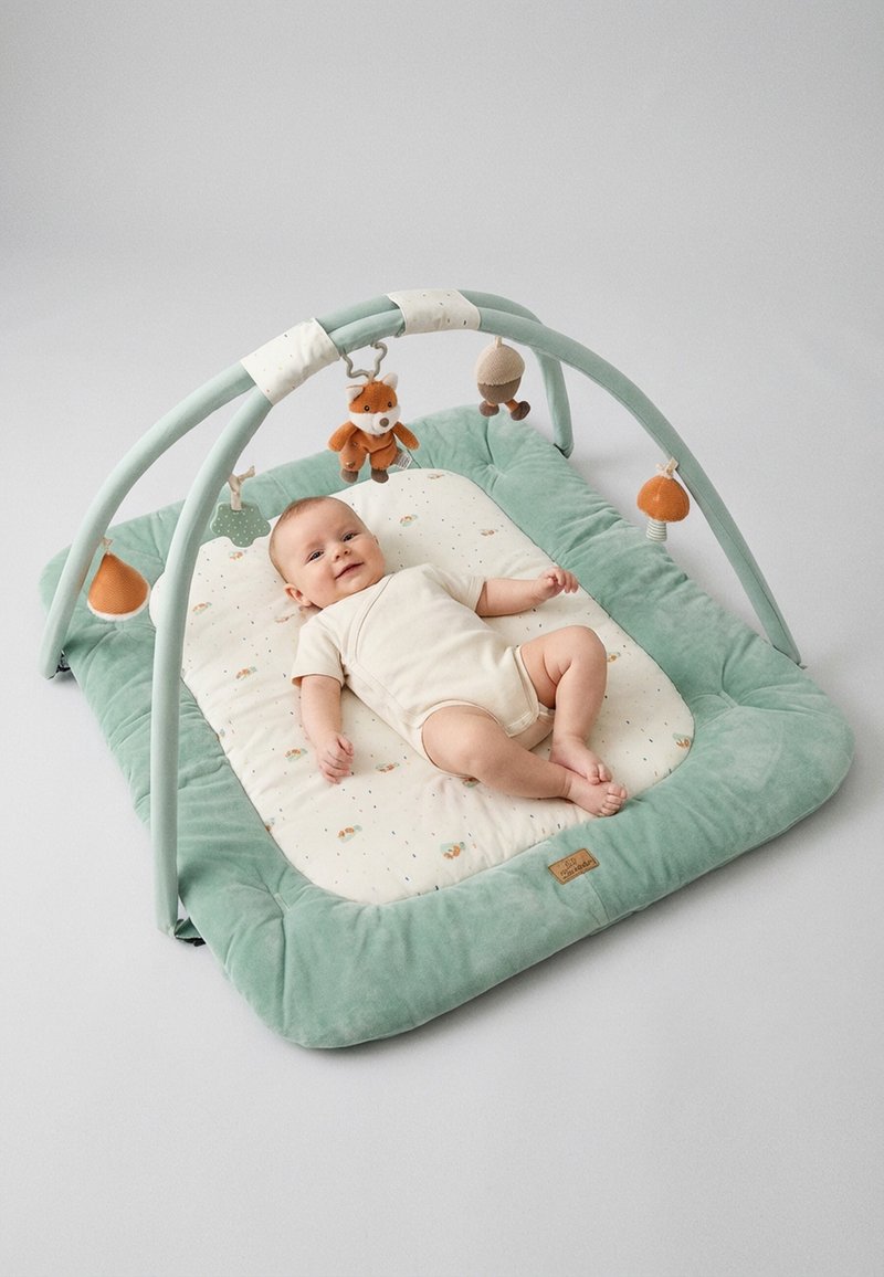 Smiling baby lying on a green and beige padded play mat with soft hanging toys, including a fox and acorn shapes.