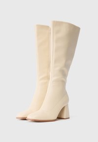 Des bottes crème de hauteur genou avec des talons carrés et des zips latéraux, présentées debout contre un fond blanc uni.