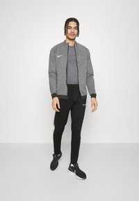 Graue Sportjacke mit schwarzen Akzenten, kombiniert mit einem schwarzen Trikot und einer Hose. Schwarze Sneakers mit weißen Details. Schlichtes, funktionales Design.