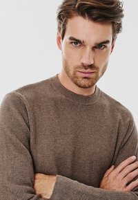 Brązowy sweter z teksturowanej dzianiny, z okrągłym dekoltem i długimi rękawami, posiadający prążkowany dekolt i mankiety. Modelka pozuje z założonymi rękami.