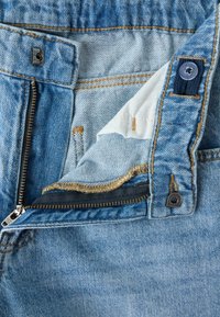 Μπλε τζιν από denim με ξεκουμπωτό φερμουάρ, μερικώς ανοιχτό μπροστινό άνοιγμα που δείχνει λευκή εσωτερική επένδυση και κουμπί σε ρυθμιζόμενο ταμπ για τη μέση.