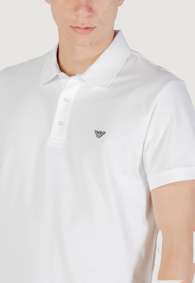 Polo shirt blanc en coton, avec un col classique, une patte de boutonnage à trois boutons et un logo brodé noir sur la poitrine gauche.