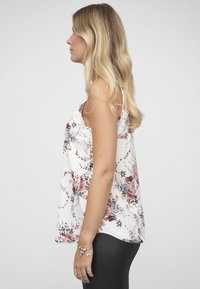 Witte tanktop met bloemmotief in roze en blauw. Heeft een delicate kanten rand op de schouders. Gecombineerd met zwarte legging.