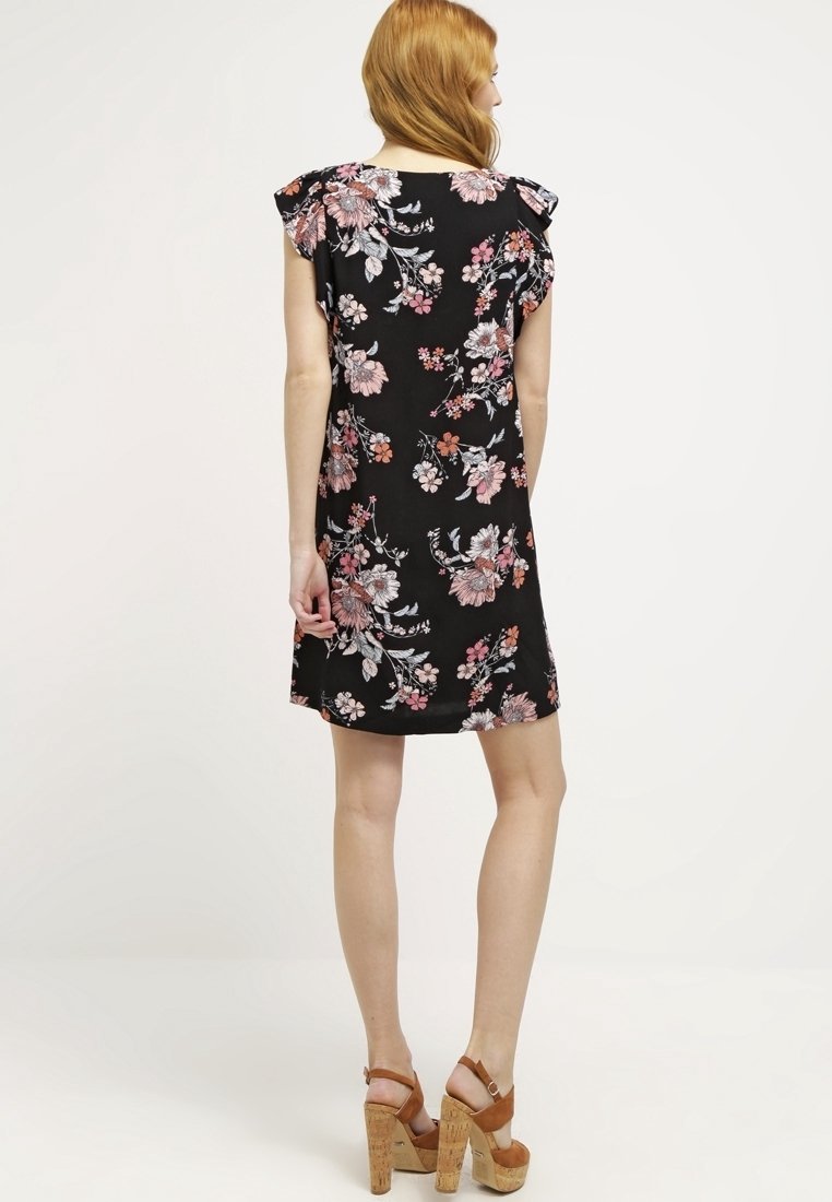 Robe noire à fleurs avec manches courtes à volants, présentant des fleurs roses et blanches. Portée avec des sandales compensées couleur tan.