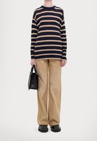 Person in einem navy-blauen und beige gestreiften Langarmshirt, beigefarbenen weit geschnittenen Hosen, schwarzen klobigen Schuhen, der eine schwarze Handtasche an der Seite hält.