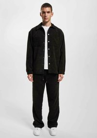 Sort kordfløjsjakke med trykknapper, to brystlommer, parret med matchende bukser, båret over en hvid t-shirt og hvide sneakers.
