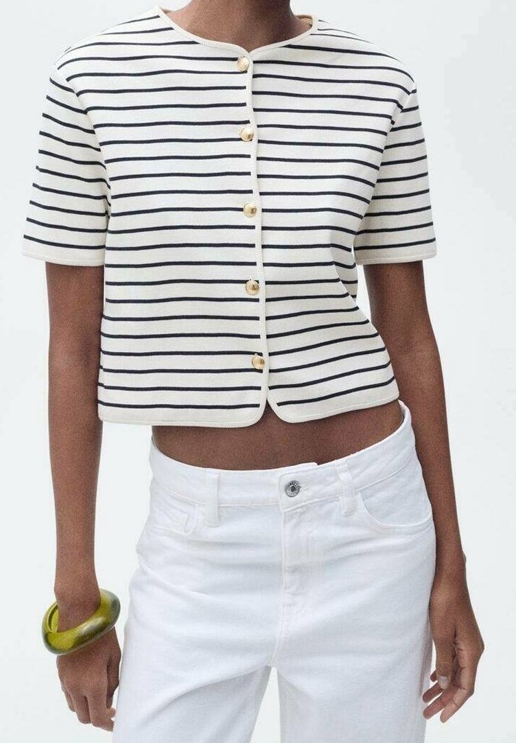 Personne portant une chemise courte blanche avec des rayures noires et des boutons dorés, un pantalon blanc taille haute et un bracelet vert au poignet gauche.