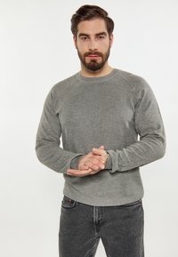 Szary sweter z okrągłym dekoltem wykonany z fakturowanej tkaniny, z rękawami raglanowymi i ściągaczami, noszony z ciemnoszarymi dżinsami. Ręce złożone z przodu.
