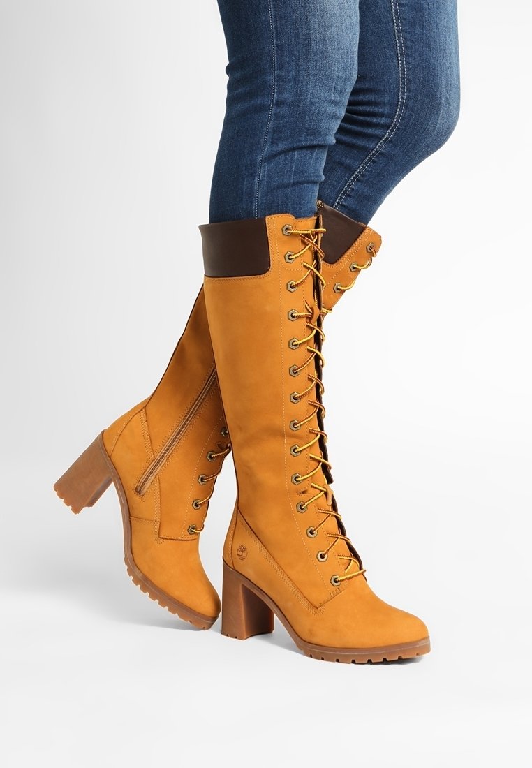 Botas de cordones de color marrón claro hasta la rodilla, con un puño de cuero marrón, tacón cuadrado, suela texturizada y ojales dorados para los cordones. Incluye detalle de cremallera lateral.