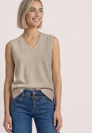 Femme souriante aux cheveux blonds courts portant un pull beige sans manches à col en V et un jean bleu sur un fond clair uni.