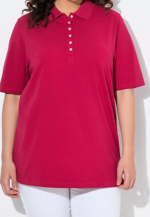 Femme portant un polo manches courtes rose foncé avec six boutons, associé à un pantalon blanc, sur un fond clair uni.