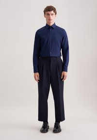 Camicia blu navy con bottoni e maniche lunghe, abbinata a pantaloni scuri a vita alta e gamba larga. Scarpe nere completano l'outfit.