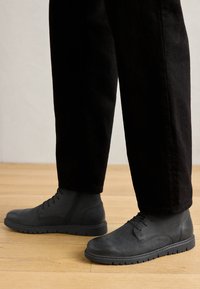 Botas de cuero negro hasta el tobillo con un acabado suave, diseño de cordones y suela de goma texturizada, combinadas con pantalones negros sobre un suelo de madera.