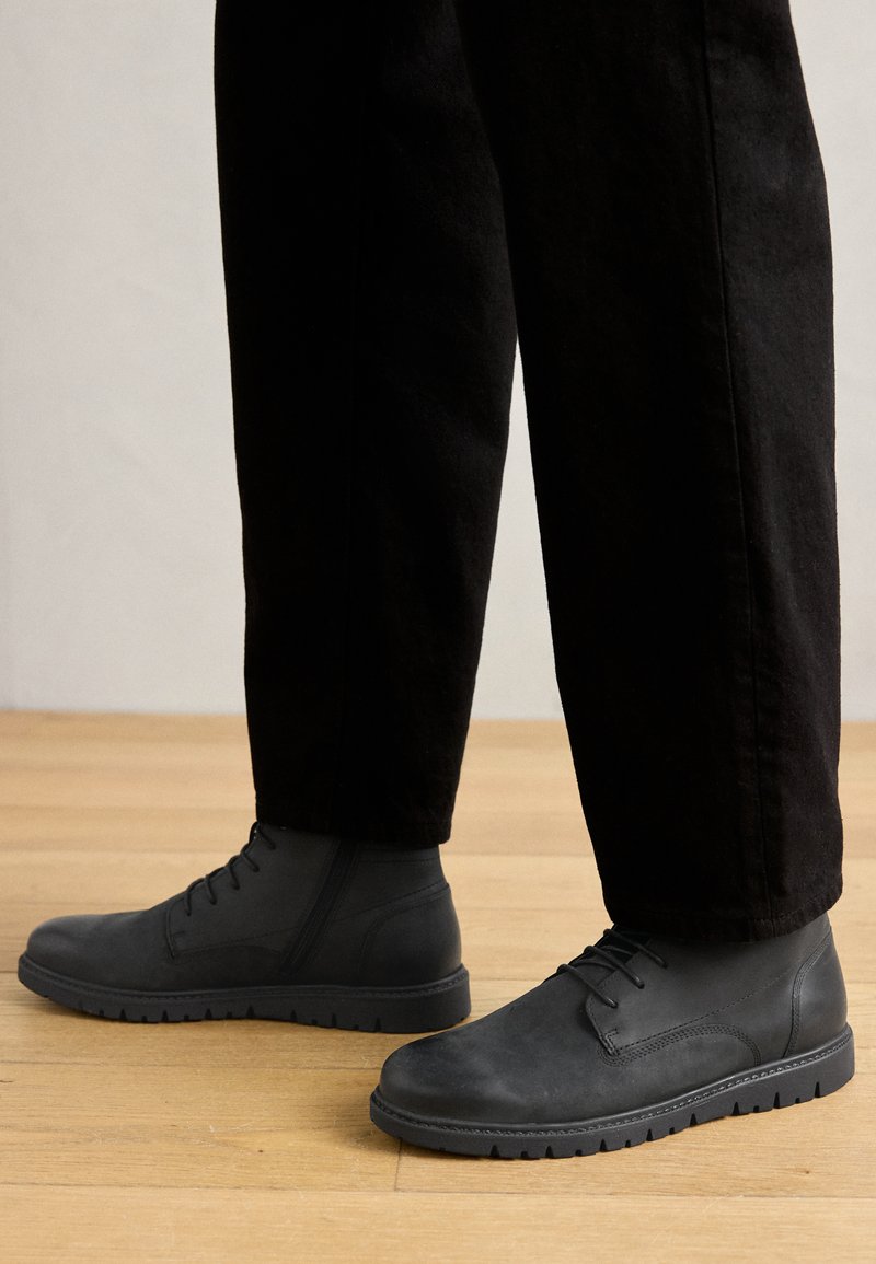 Stivaletti neri in pelle con una finitura liscia, design con lacci e suola in gomma texturizzata, abbinati a pantaloni neri su un pavimento di legno.