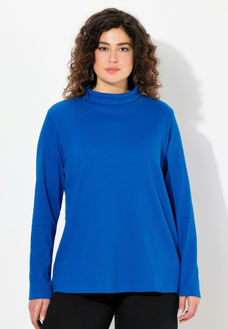 Maglietta a maniche lunghe blu con collo alto in un tessuto liscio e leggero. Presenta una vestibilità rilassata e un collo alto. Abbinata a pantaloni neri.