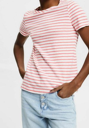 Persoon draagt een wit overhemd met korte mouwen met roze horizontale strepen en lichtblauwe denim spijkerbroek met knopen, handen gedeeltelijk in de zakken.