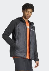 Veste isolée noir et gris pour homme avec capuche, présentant un design matelassé et des poignets élastiques, portée sur une chemise orange.