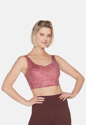 Frau mit kurzem blondem Haar, die einen gemusterten pinkfarbenen Sport-BH und dunkle, hoch taillierte Leggings trägt, steht mit den Händen in den Hüften.