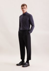 Camicia scura aderente con colletto a punta, abbinata a pantaloni neri a vita alta. Scarpe e calzini neri completano il look. Tessuto dalla texture liscia.