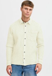 Camisa texturizada de un beige claro con diseño de botones, dos bolsillos en el pecho, cuello redondo y botones grises. Mangas largas con puños.
