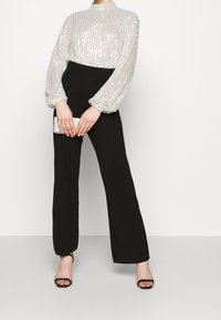Blusa di paillettes argento lucido con maniche lunghe e gonfie abbinata a pantaloni neri a vita alta e a gamba larga. Modella tiene una pochette luccicante.